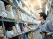 Young man worker doing stocktaking of product in cardboard box on shelves in warehouse by using digital tablet and pen.
