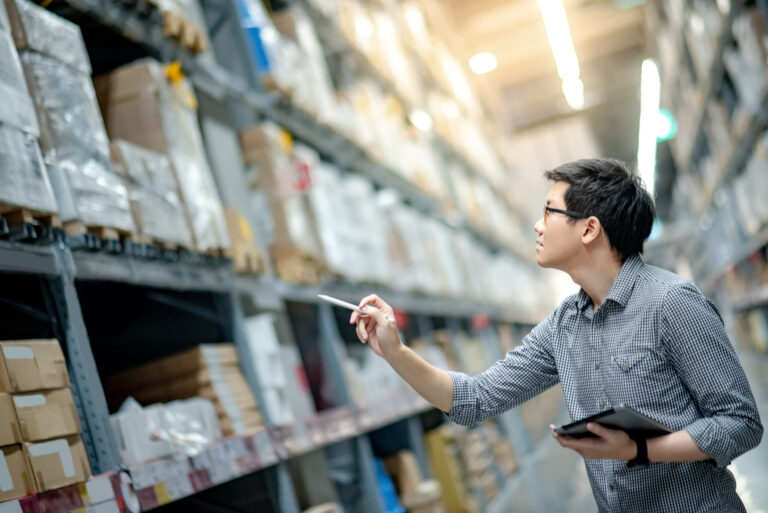 Asian man doing stocktaking by using tablet in warehouse Young man worker doing stocktaking of product in cardboard box on shelves in warehouse by using digital tablet and pen.