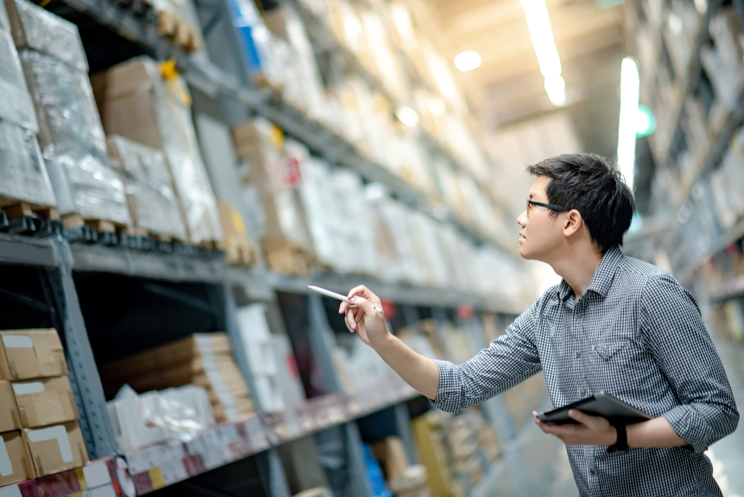Young man worker doing stocktaking of product in cardboard box on shelves in warehouse by using digital tablet and pen.